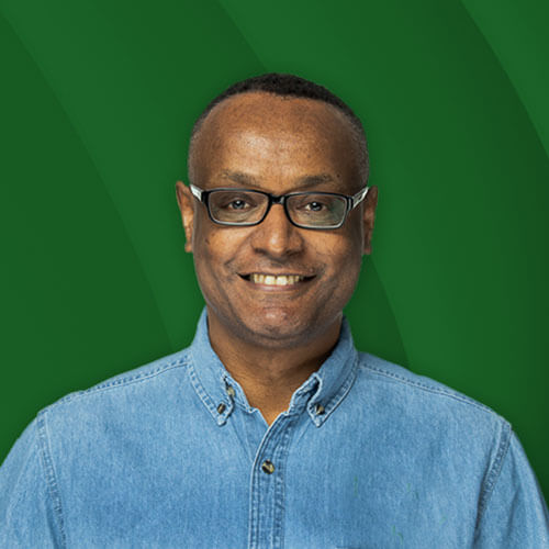 A headshot of Teshome Jiru. He is smiling into the camera. He is wearing glasses and a blue denim button down shirt.