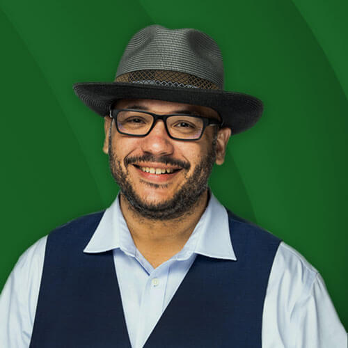 A headshot of David Burchfield, who is smiling into the camera. He is wearing a fedora hat, glasses, a dark blue vest and a light blue button down shirt.