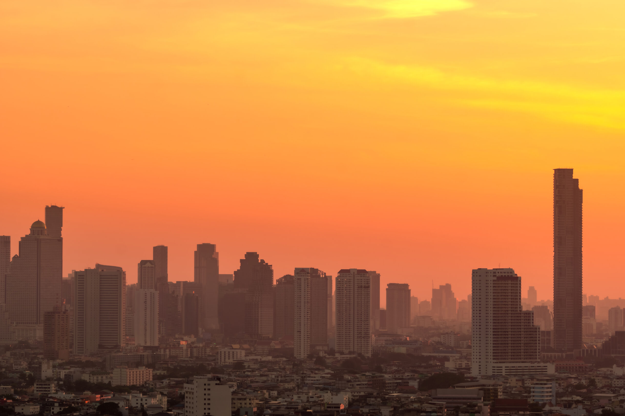A city scape of small and tall buildings is shrouded in a cloak of smog at sunset. The color of the smog is deep orange on the lower left to yellow in the upper right.
