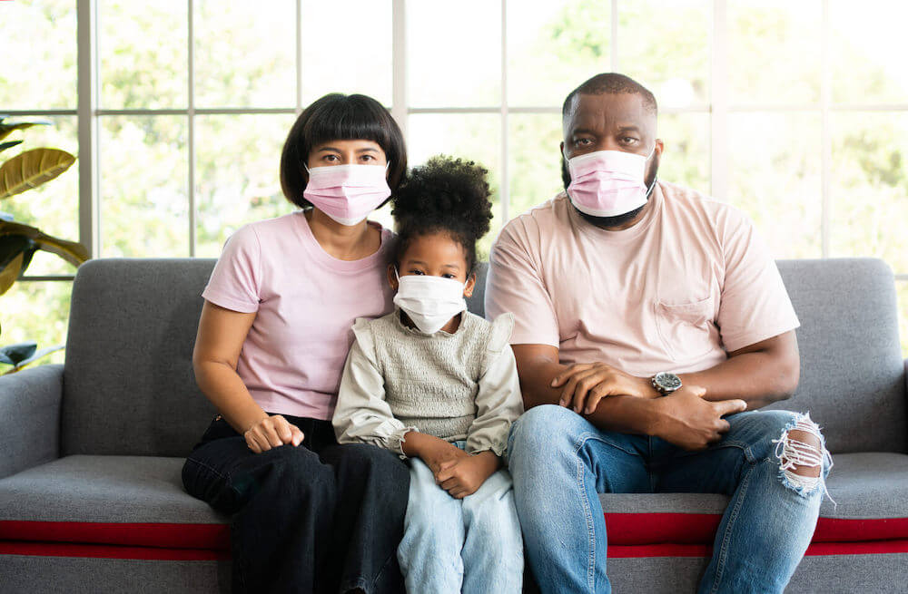 People presenting as a heterosexual mixed race couple and their female-bodied child are sitting on a couch wearing masks and looking very serious.