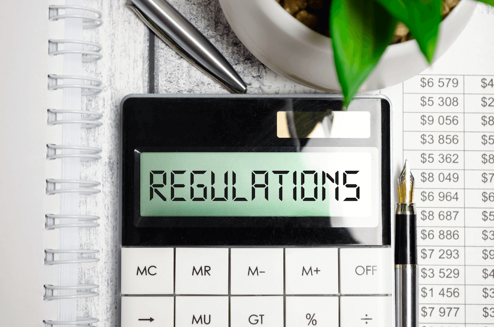 A image of a calculator with the word "REGULATIONS" where numbers would be is surrounded by the edge of a binder, some pens, a plant, and a table with different dollars amounts in each cell.