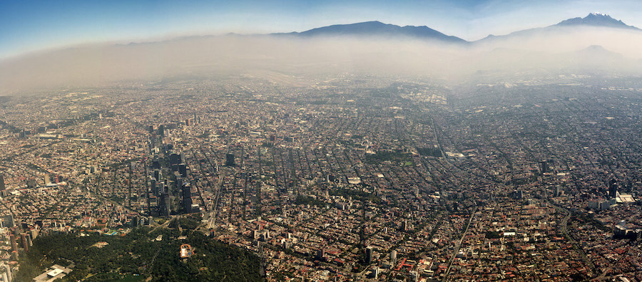 A picture of Mexico City from overhead. Smog covers the city and almost blocks out the mountains behind entirely.