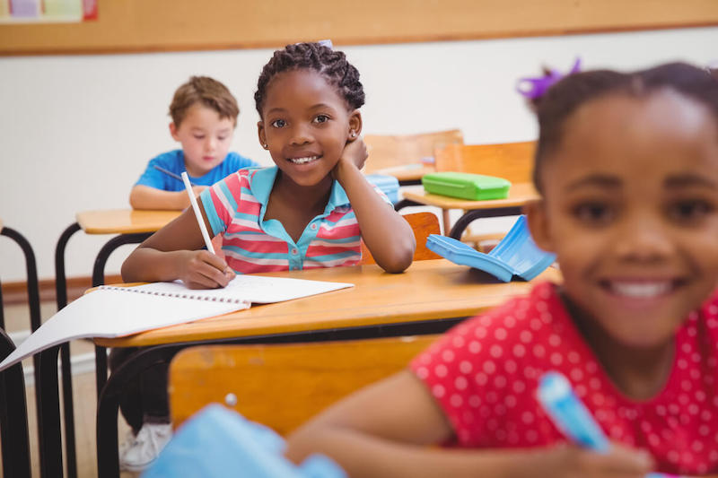 A picture of 3 kids in school. A very young child who presents as a Black girl is in focus in the middle of this photo, smiling and looking right into the camera while holding a pencil over a notebook at a school desk. In the foreground is a blurry person who presents as a very young Black girl and is also smiling, looking into the camera and holding a pen. In the background, a person who presents as a young white boy is looking down at the desk.