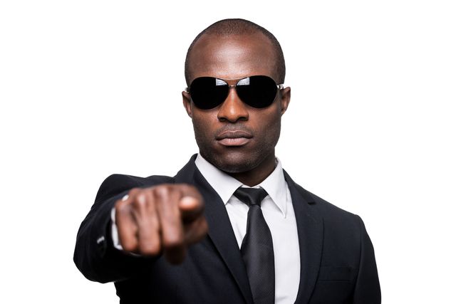 A person presenting as a Black male is pointing and looking directly into the camera. They are wearing sunglasses and have a serious expression on their face.