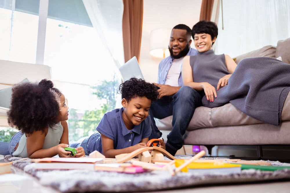 A family presenting as mixed race (Black and African American Father and an Asian Mother) lounge on the couch. Two children play on the carpet of a modern apartment. Everyone is smiling. The Father is holding a computer and one of the children is looking up at the couch they're on.