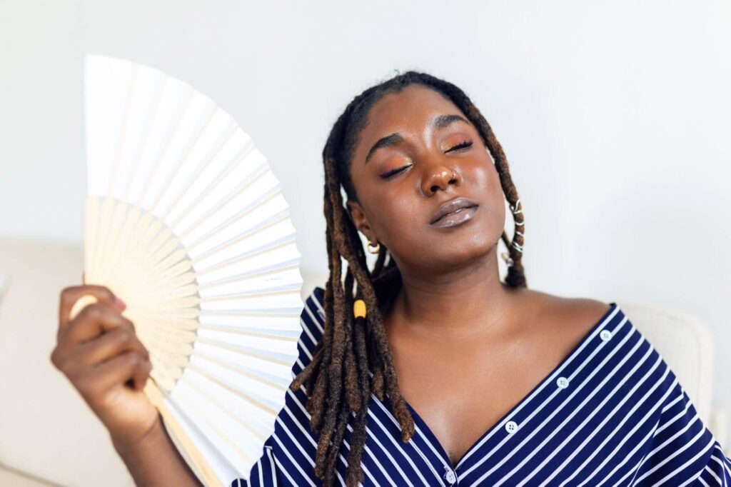 A person presenting as a Black female bodied person is fanning themselves. They look kind of sleepy and their eyes are closed, as if they are feeling very hot.