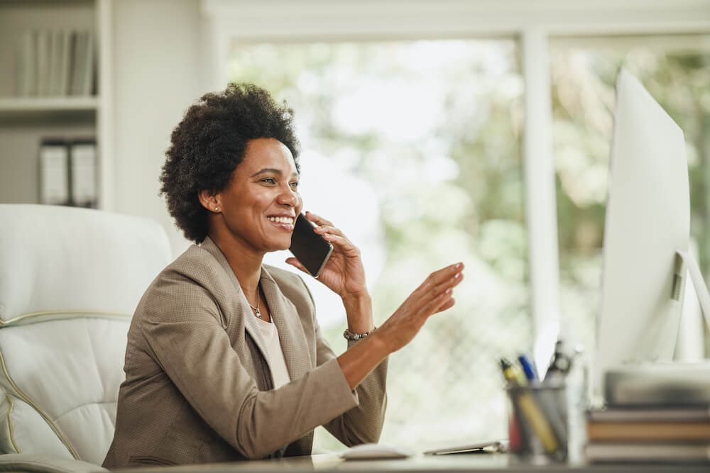 A person presenting as a Black woman or assigned-female-at-birth is on a cell phone at their desk. They are smiling and holding up their hand in an animated conversation. They are surrounded by white furniture. Windows behind them give way to greenery.