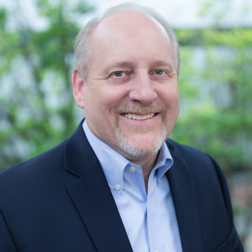 A picture of Larry Bekkedahl, who appears to be a white man with gray hair, wearing a dark blue business jacket over a light blue button down shirt. Green vegetation is in the background.